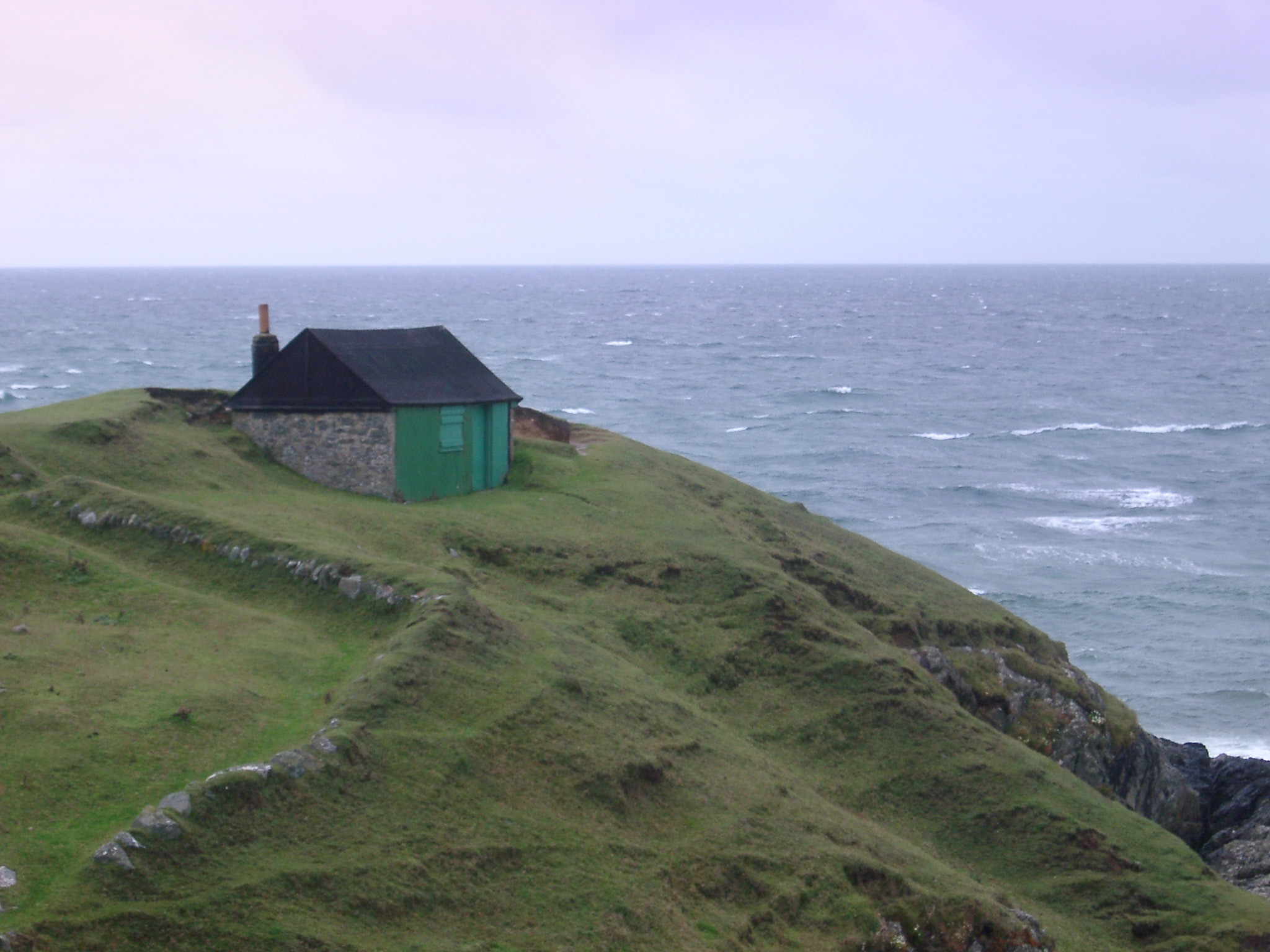 an image of Stormy coastal landscape with the roof of a small hut or cottage on a lush promontory with pastures overlooking a wild ocean with breaking waves