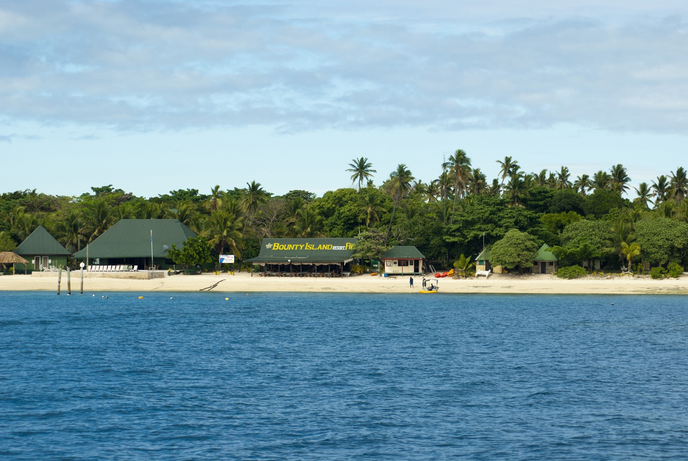 an image of Bounty Island resort, Fiji, on an idyllic tropical island with tourist visible on the sandy beach in front of the buildings