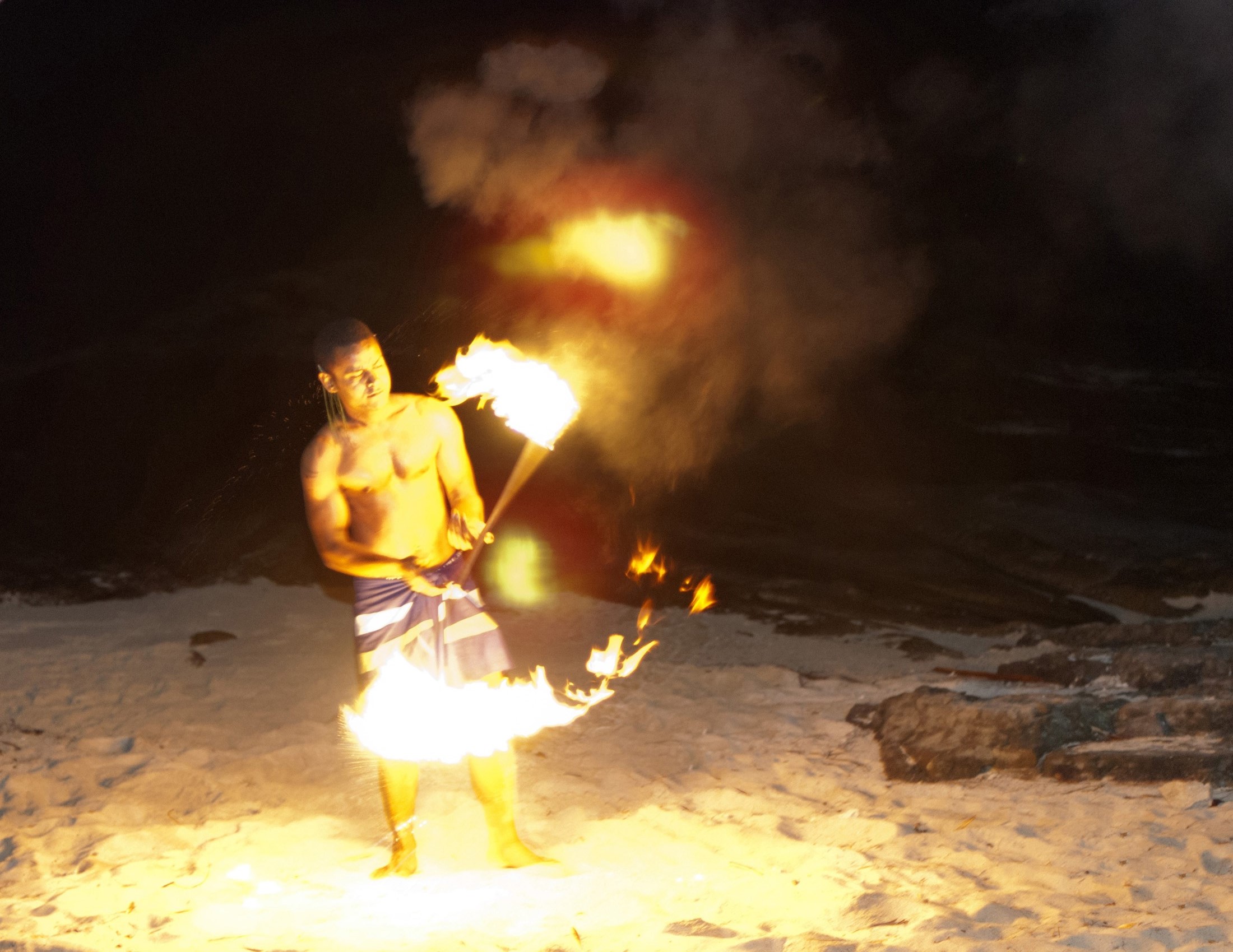 an image of Shirtless barefoot fire dancer on Fiji twirling flaming sticks during a night performance on a sandy beach