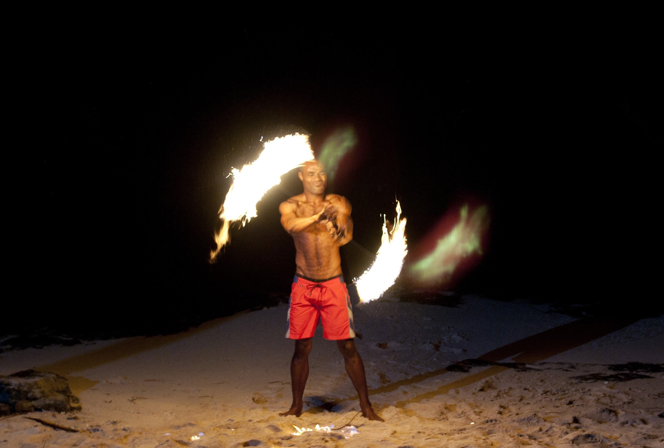 an image of Fijian fire dancer performing with flaming branches on a sandy beach twirling them around his naked torso