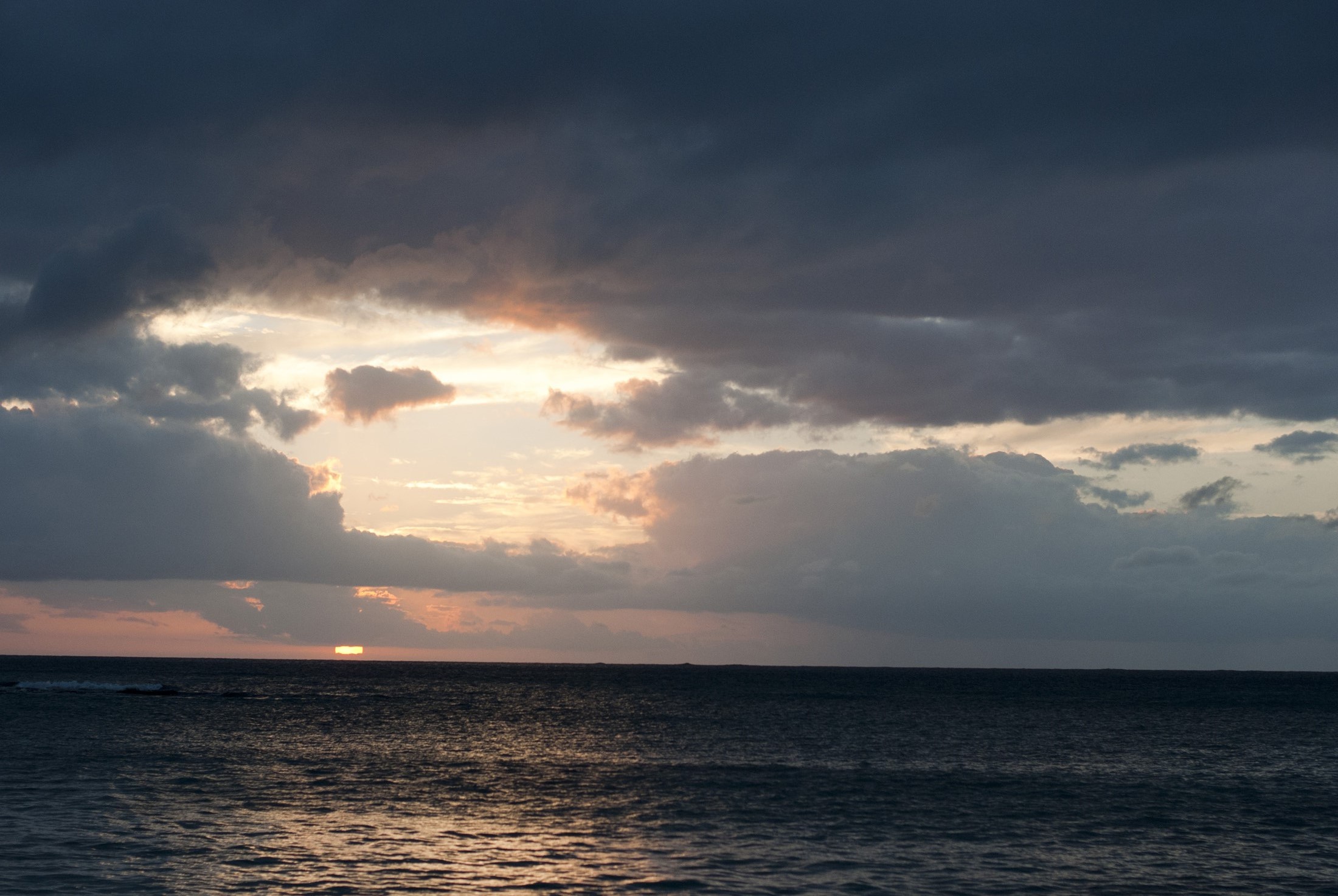 an image of Setting sun over the ocean shing through a break in the heavy cloud cover with a delicate pink glow illuminating the water