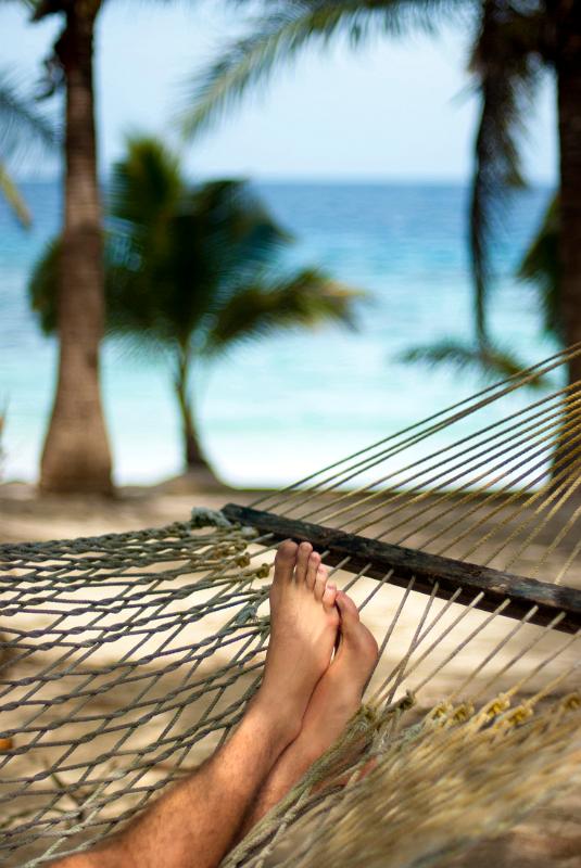 Cropped view image of male feet relaxing in a hammock overlooking a tropical beach with palm trees, shallow dof