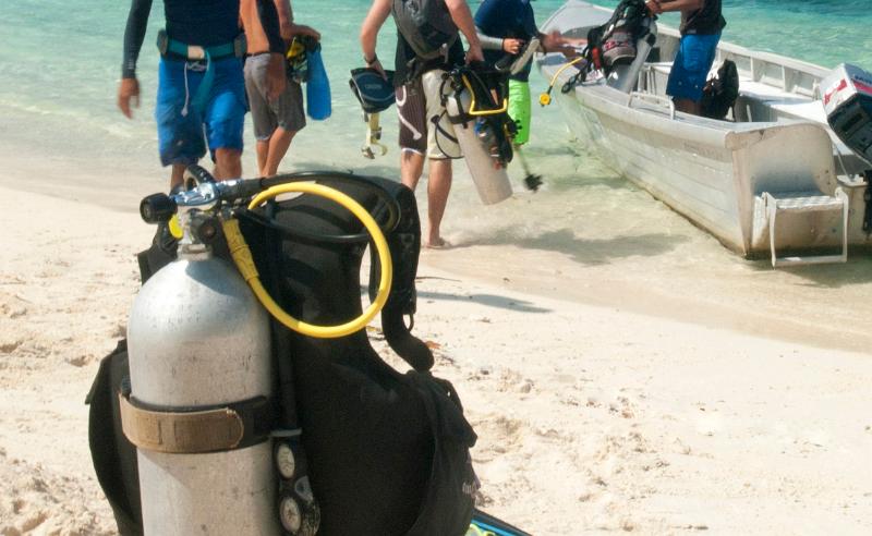 An air tank and diving equipment wait on a sandy beach as a group of divers load their boat before going for a scuba dive