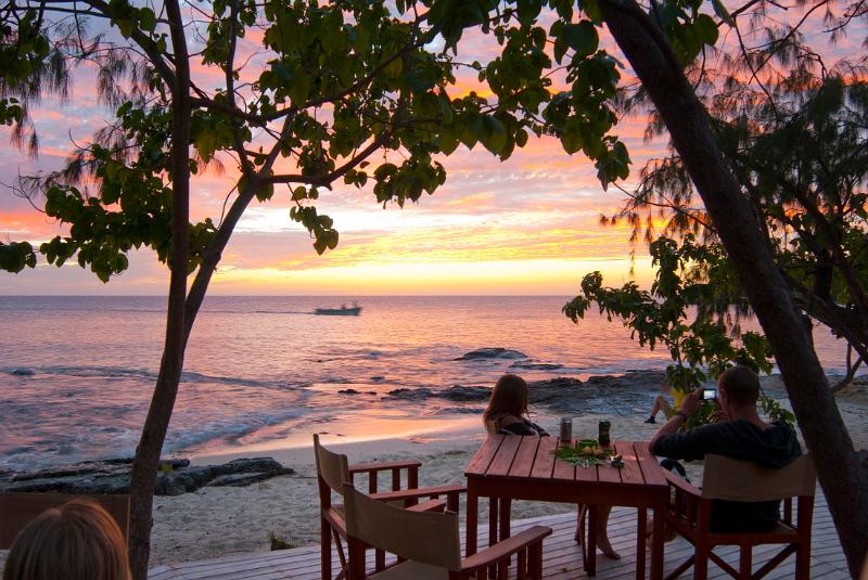 People seated at a table on a deck overlooking the beach and the ocean enjoying a beautiful tropical sunset