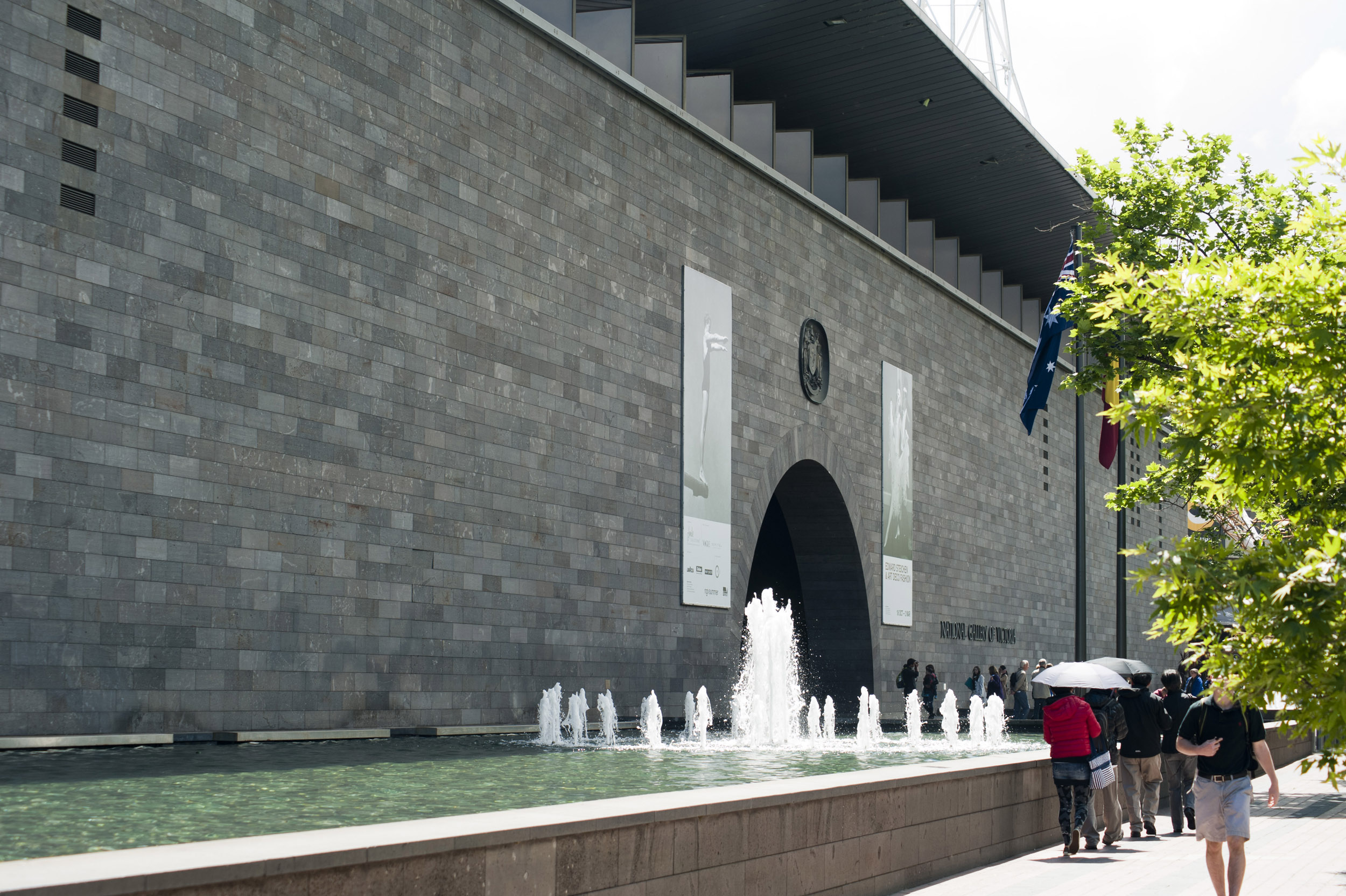 an image of People outside the Melbourne National Gallery, Australia walking alongside the ornamental fountains and pond under umbrellas on a hot sunny day