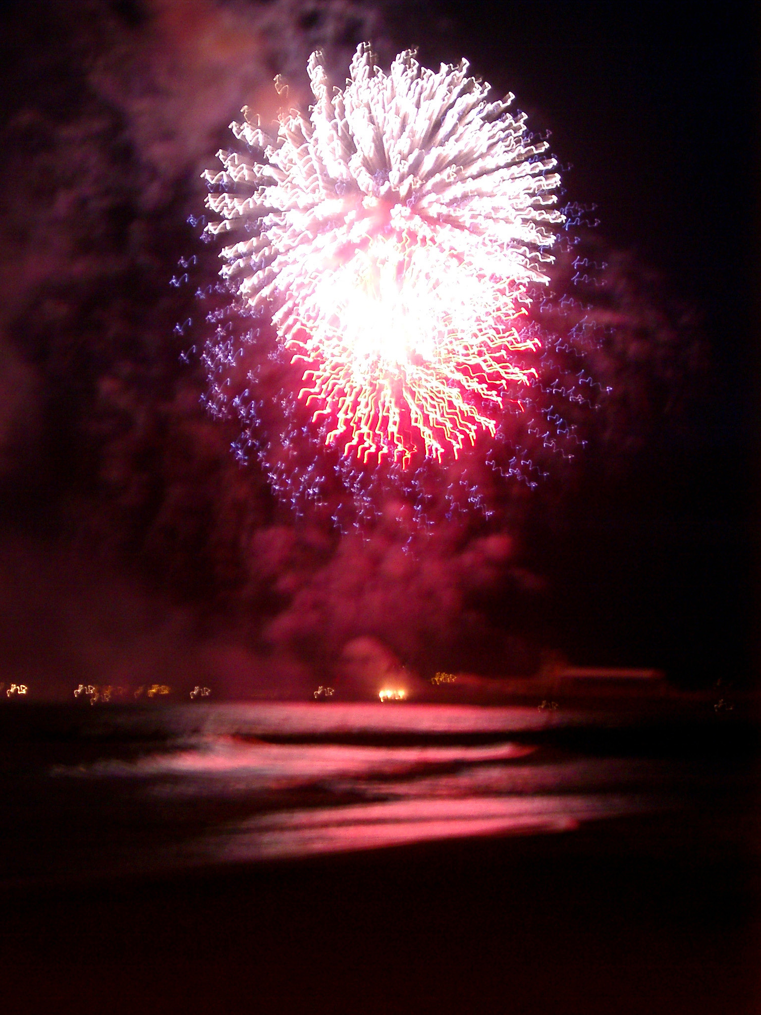 an image of Long Exposure of Pink Fireworks Exploding in Sky Above Water at Night