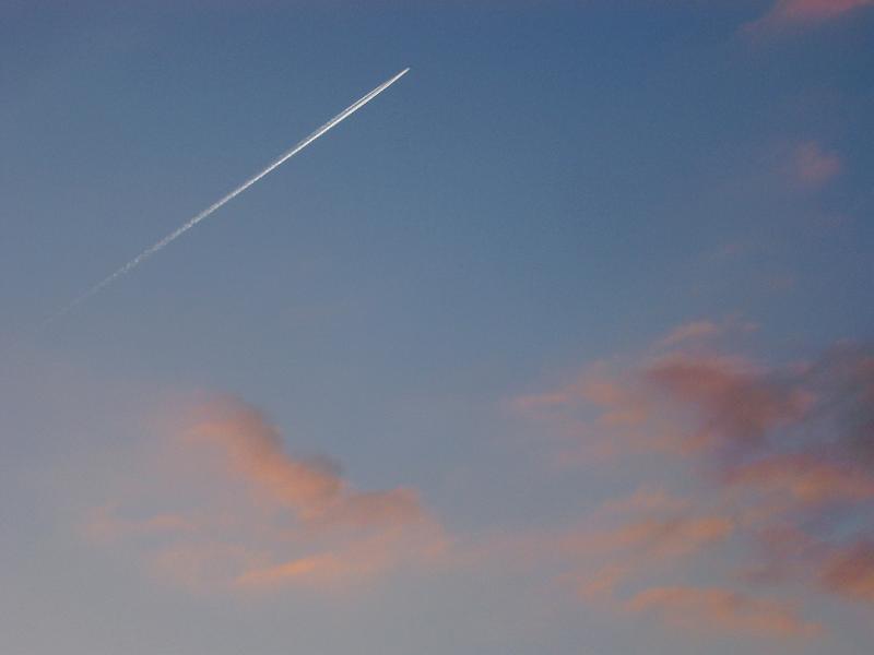 Jet trail or contrail formed by condensation of vapor and ice particles streaking through a blue sky at sunset with colorful pink clouds