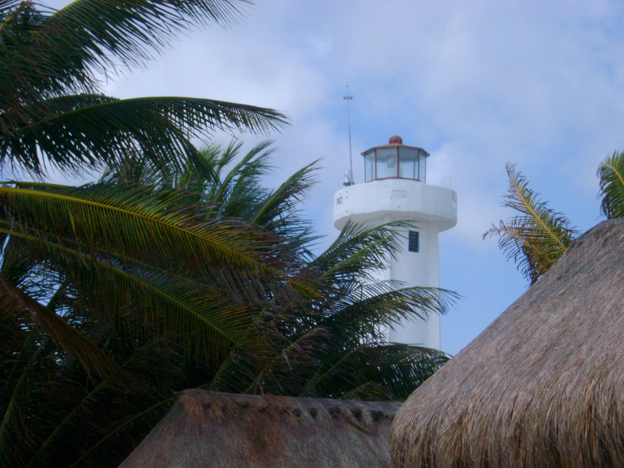 an image of Lighthouse surrounded with palm trees viewed over the thatched roofs of beach cottages in Mexico