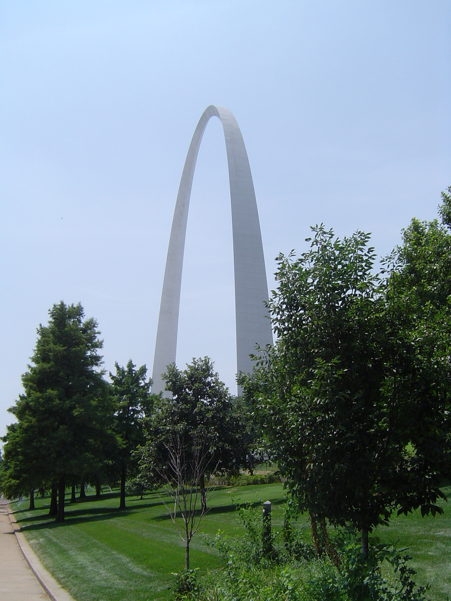 an image of Gateway Arch - Famous St Louis City Symbol, Surrounded by Green Trees. Isolated on Light Blue Sky Background.