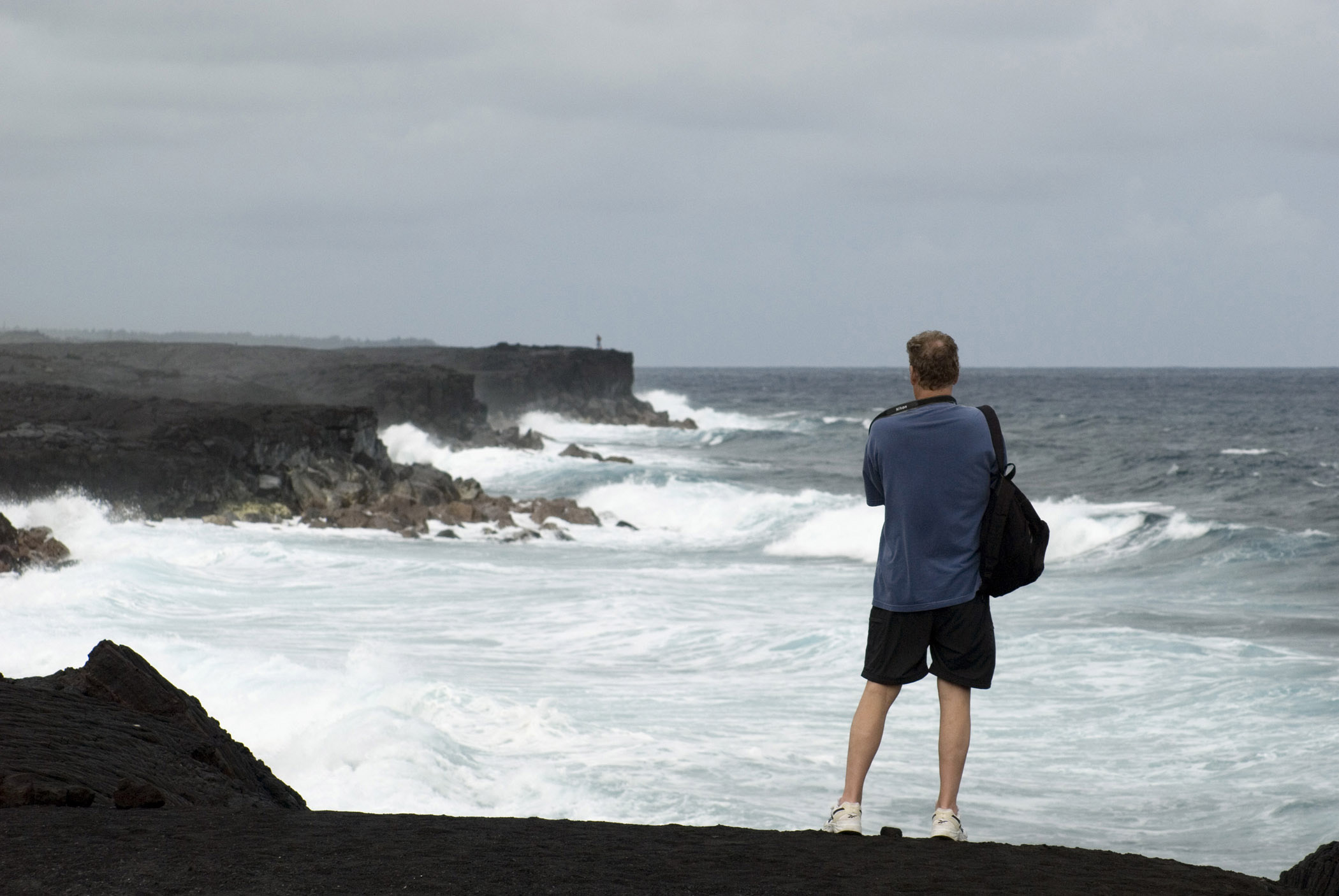 an image of Tourist standing with his back to the camera on the black volcanic sand on Kalapana beach, Hawaii looking out over the ocean waves and rocks