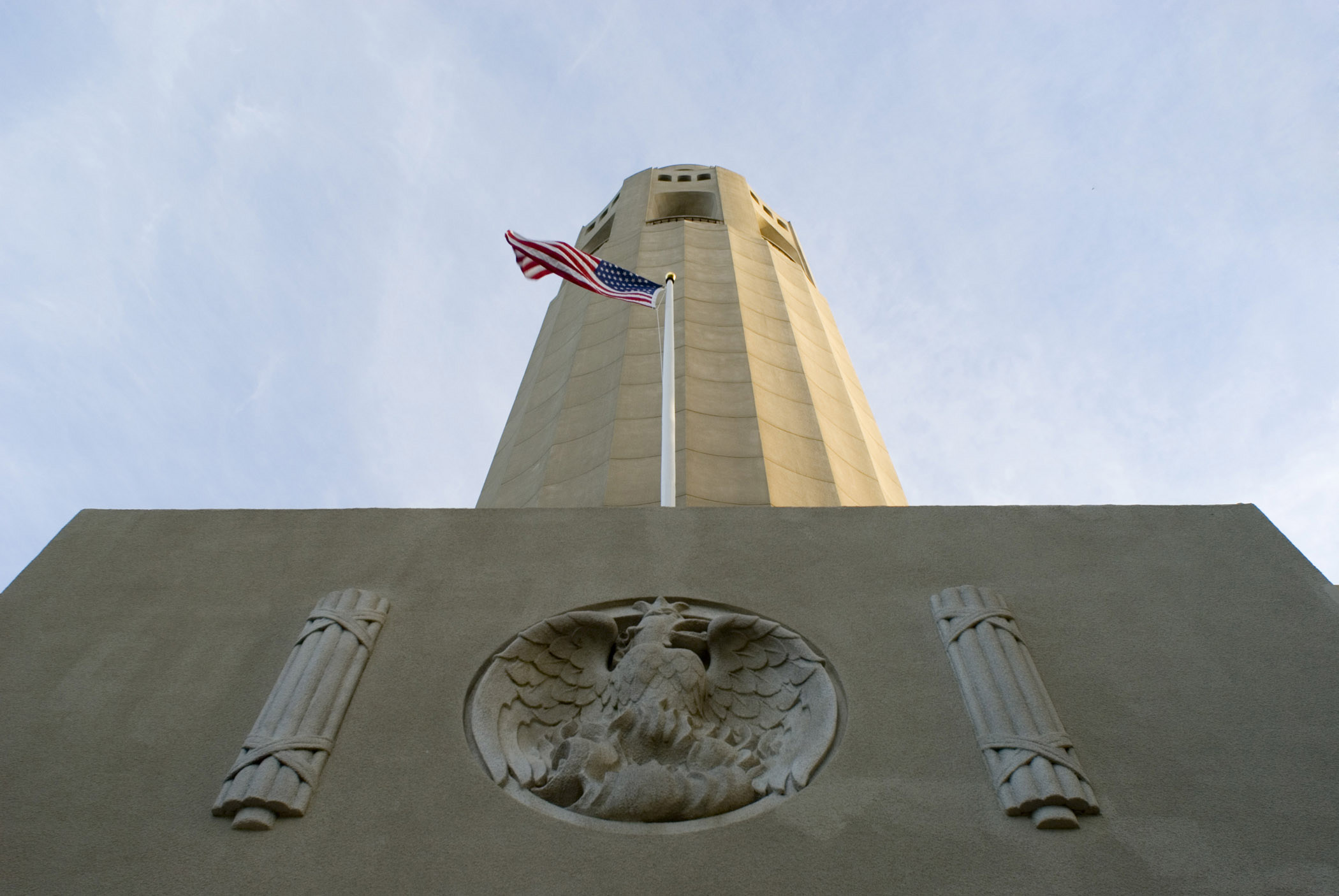 an image of Famous Old Tall Coit Tower Structure with American Flag in Worms Eye View. Located in San Francisco. Isolated on Very Light Blue Sky Background.