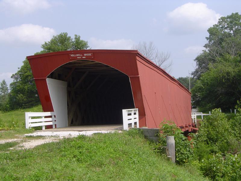 Close up Covered Bridge Surrounded by Green Grasses and Tall Trees on Lighter Blue Sky Background.