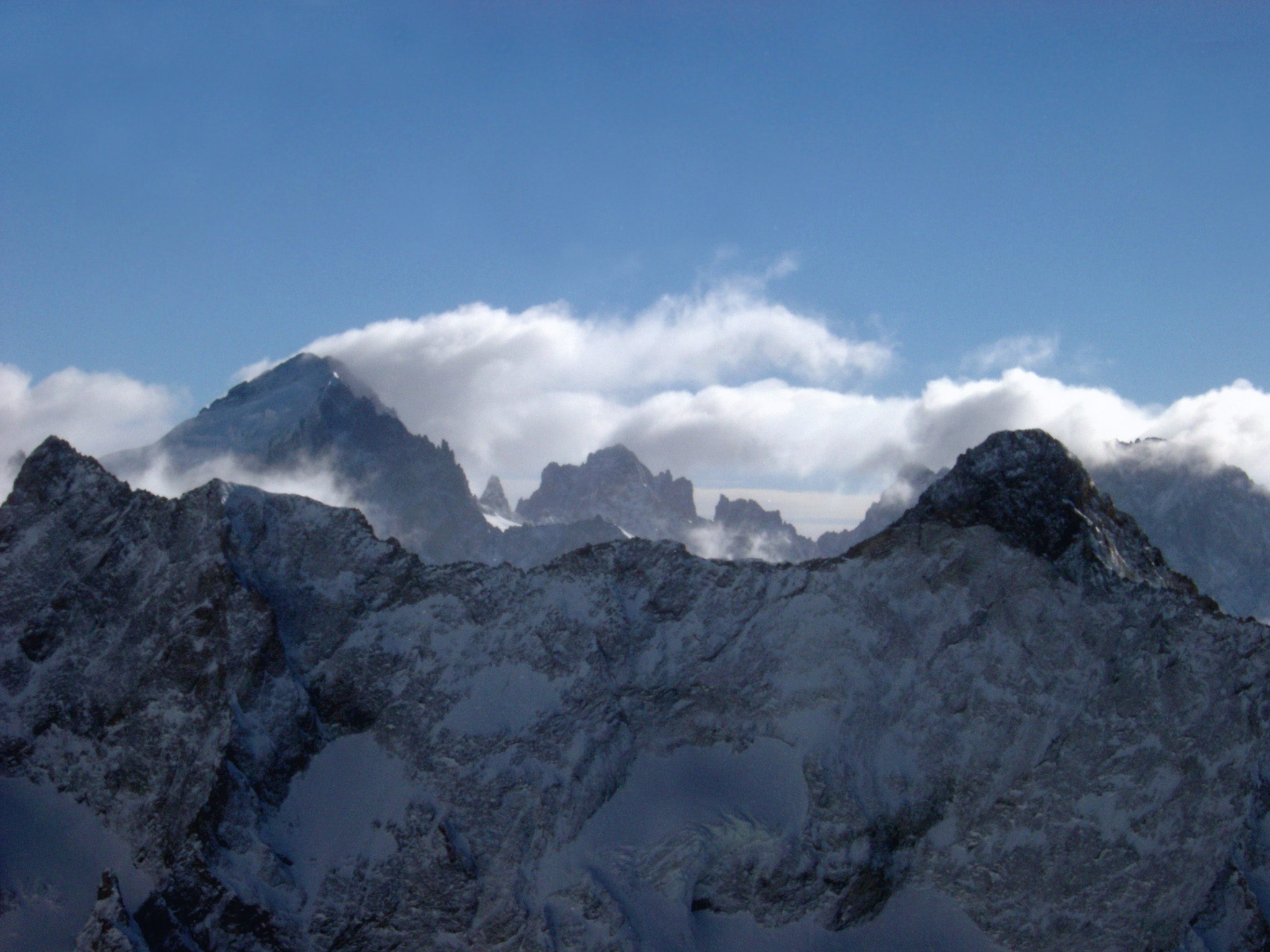 an image of Alpine winter holiday with a view over rugged snow covered mountain ranges and peaks with fluffy white cloud cover under a clear blue sunny sky